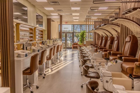 Modern nail salon interior with beige workstations, pedicure chairs, and a reception desk illuminated by natural and overhead lighting
