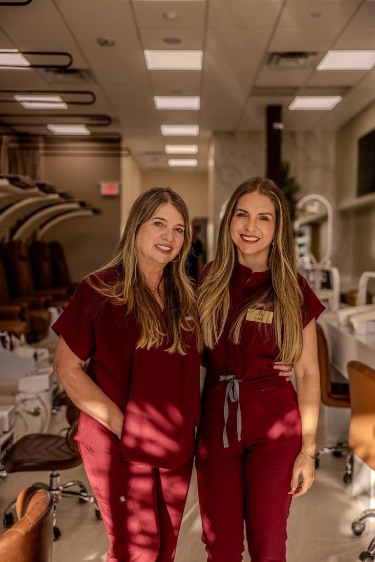 Two women in burgundy scrubs smiling in a medical or salon treatment room with white cabinets and ceiling lights