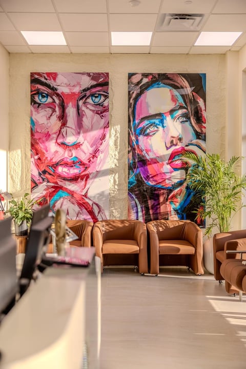 Modern office lobby with colorful abstract face paintings on walls, brown leather chairs, green plants, and natural light from ceiling panels
