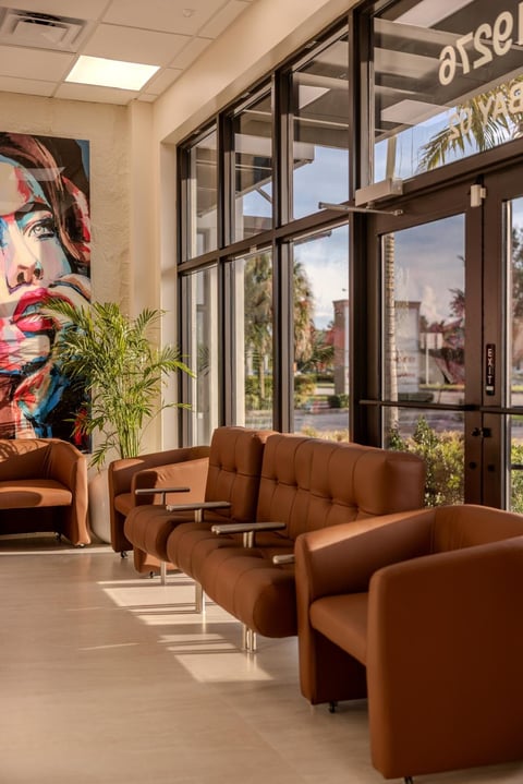 Modern office lounge with brown leather armchairs, floor-to-ceiling windows overlooking city view, artwork on wall, and green plants.