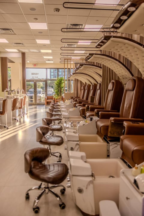 Modern nail salon interior with rows of beige pedicure stations featuring brown leather chairs lined along a polished floor, recessed ceiling lights, and floor-to-ceiling windows.